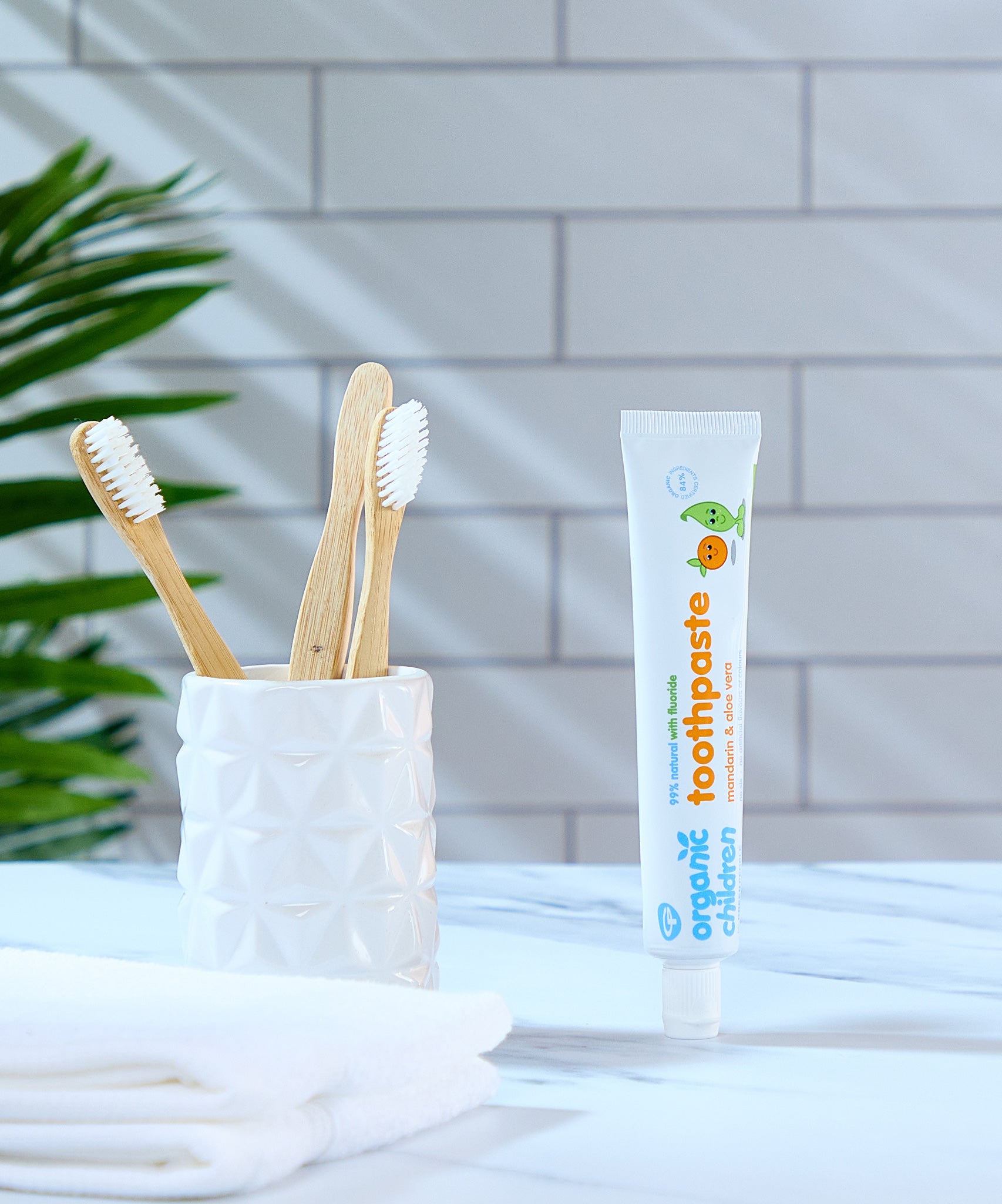 A tube of Organic Children Toothpaste Mandarin & Aloe Vera with Fluoride placed on a marble surface next to a ceramic pot containing bamboo toothbrushes. 