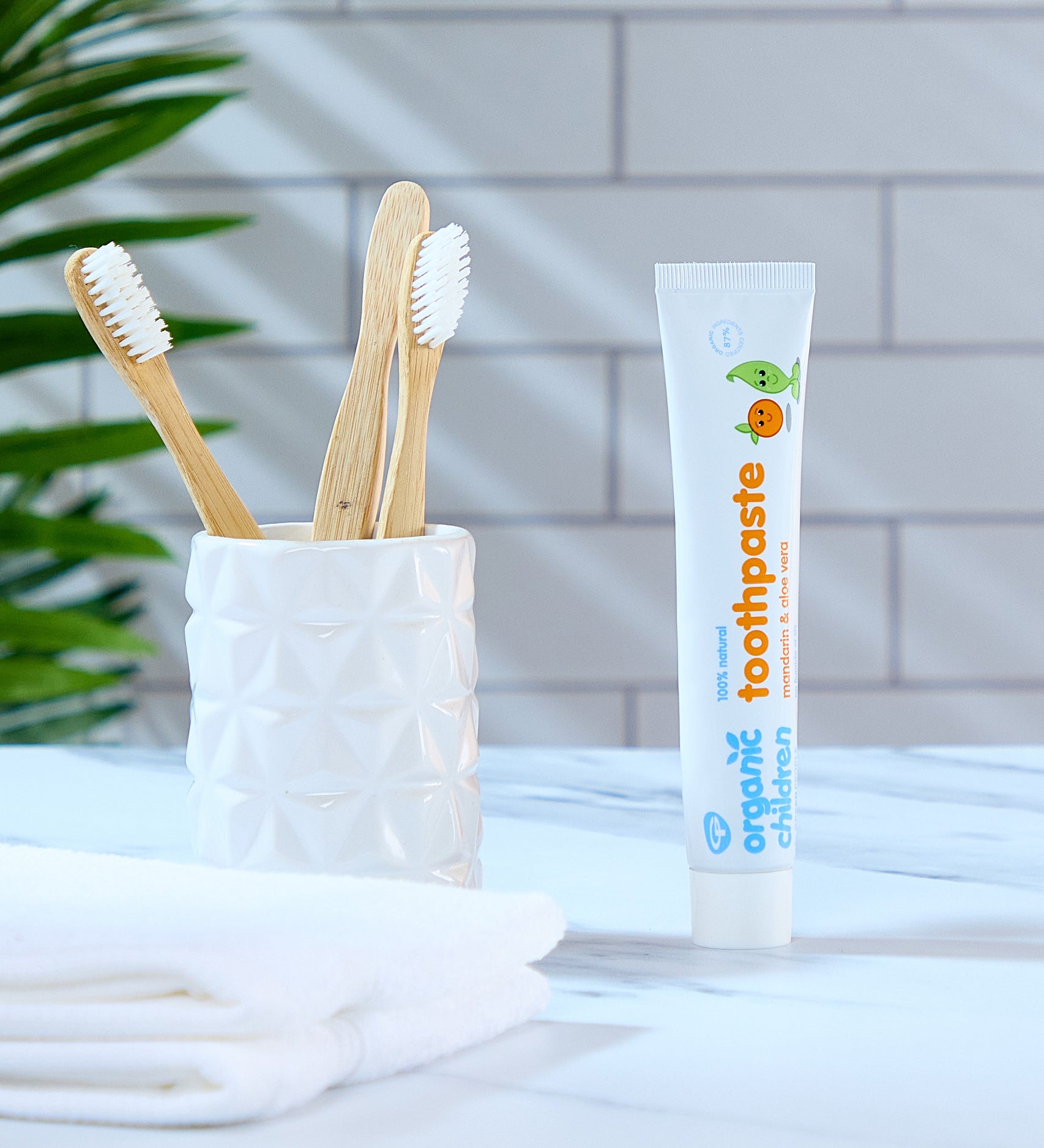 A tube of Organic Babies Toothpaste Mandarin & Aloe Vera pictured next to a pot of bamboo toothbrushes. A green leafy plant can be seen in the background. 