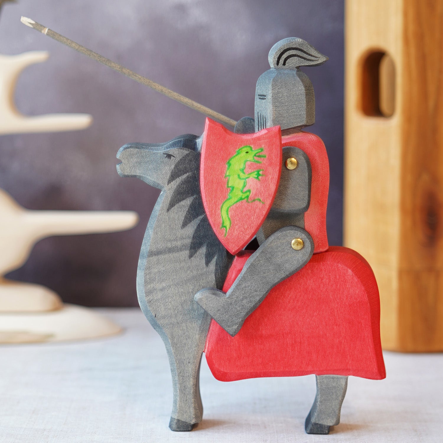 A wooden figure of a knight in black armor sitting aloft a black horse. The knight holds a shield and a jousting stick. The shield and parts of the body armor are red in colour. There is a green dragon motif on the shield.  In the background is a wooden t