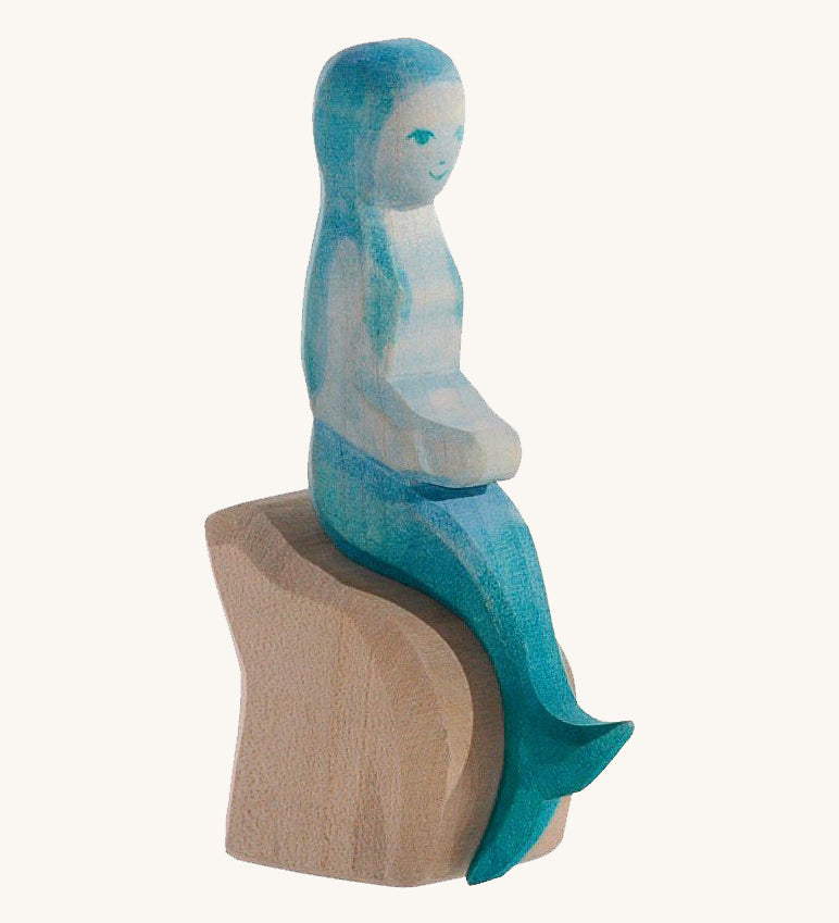 A wooden Ostheimer mermaid with blue hair sitting on a rock and on a cream background.