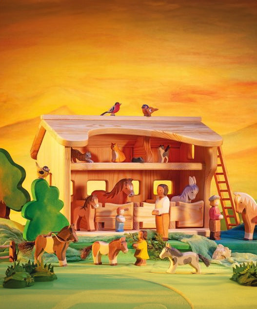 Ostheimer wooden toy Horse Stable placed in a small world play scene. The scene has lots of Ostheimer animal figures along with people and greenery and trees. 