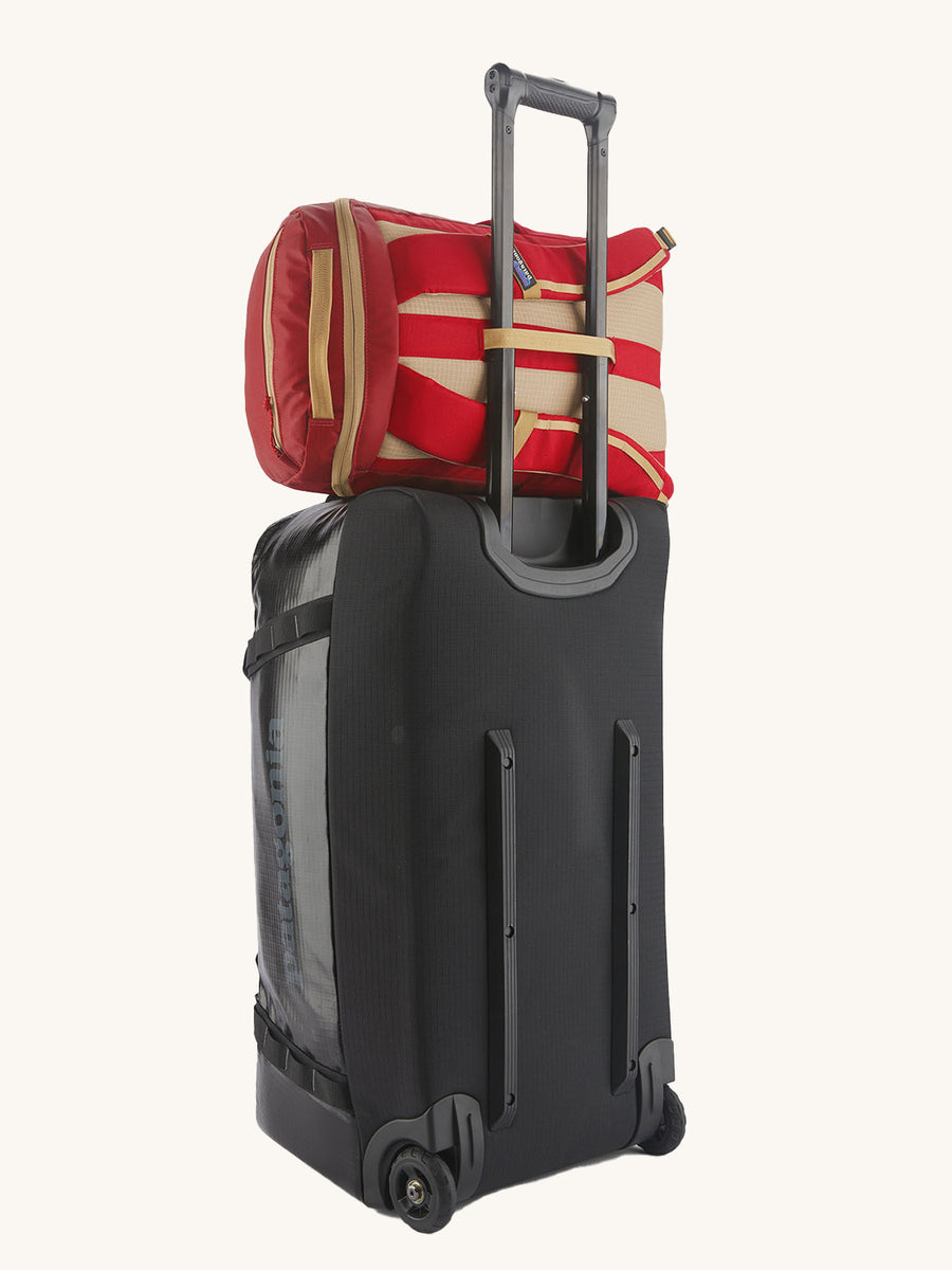 Patagonia Recycled Black Hole Backpack 25L in Touring Red hooked over the handle of a luggage roller case on a cream background 