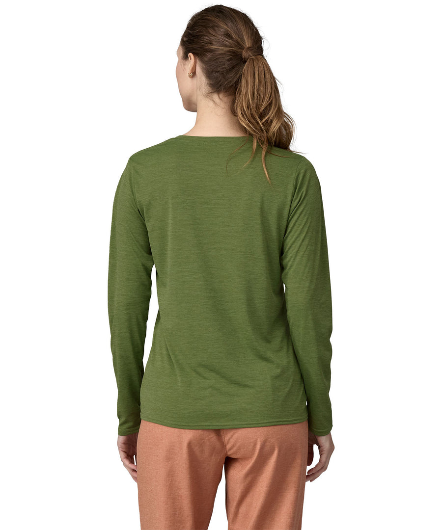 A woman wearing a Patagonia Women's long sleeved Cap Cool Daily Graphic Shirt in a Green X-Dye colour. The woman is stood in front of a plain background. The woman is facing away from the camera, showing the back of the top. 