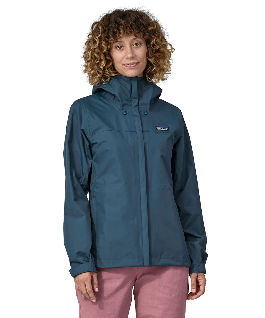 A woman wearing a Patagonia Women's Torrentshell 3L Rain Jacket in a Lagom Blue colour stood in front of a plain background. 