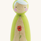 A wooden handmade Peepul peg doll wearing a light green cape with a red poppy painted on the front on cream background.