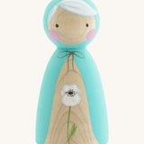 A wooden handmade Peepul peg doll wearing a light blue cape with a white poppy painted on the front on cream background.