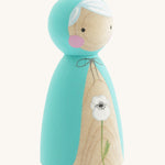 A side photo of the wooden handmade Peepul peg doll wearing a light blue cape with a white poppy painted on the front on cream background.