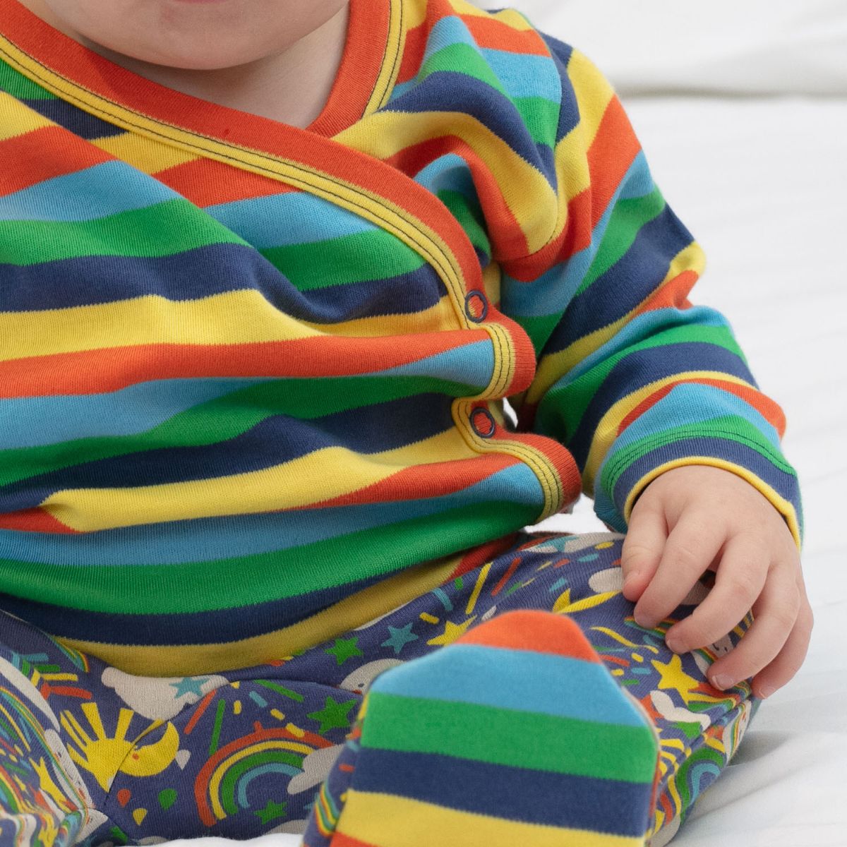 baby wearing  three piece baby set with a bright rainbows and planets design from piccalilly