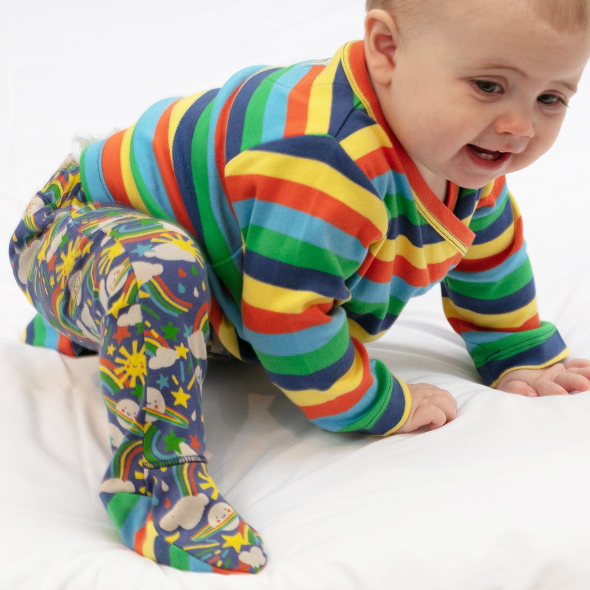 baby wearing  three piece baby set with a bright rainbows and planets design from piccalilly