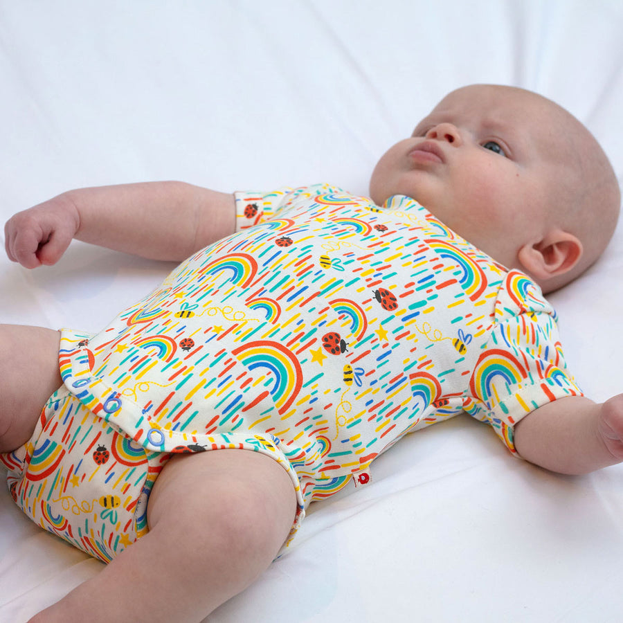 Baby laying on a white sheet wearing the Piccalilly short sleeve rainbow baby body suit