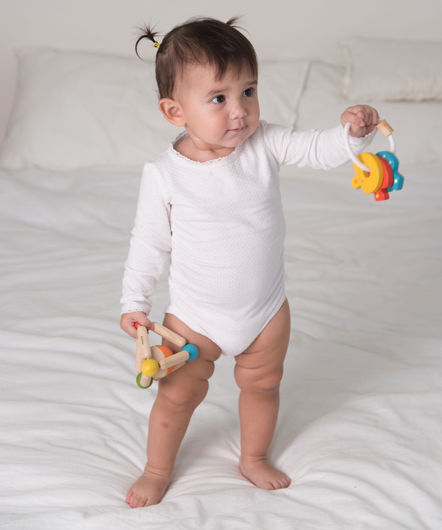 A baby standing on as bed holding onto the PlanToys Baby Key Rattle in one hand and the PlanToys Baby triangle clutching toy in the other. 

