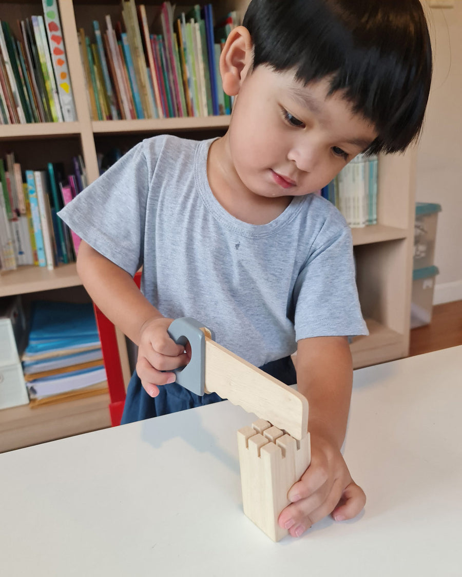 Boy playing with the plastic free Plan Toys woodwork saw on a white table in front of a large bookshelf