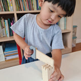 Boy playing with the plastic free Plan Toys woodwork saw on a white table in front of a large bookshelf