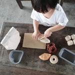 Close up of the Plan Toys wooden play food bread set laid out on a wooden bench next to its fabric storage bag and some sliced bread