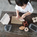 Close up of the Plan Toys wooden play food bread set laid out on a wooden bench next to its fabric storage bag and some sliced bread