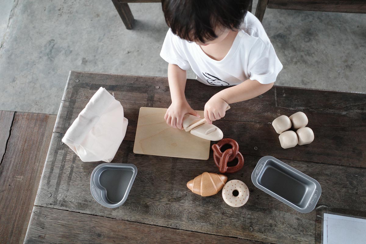 Close up of the Plan Toys wooden play food bread set laid out on a wooden bench next to its fabric storage bag and some sliced bread