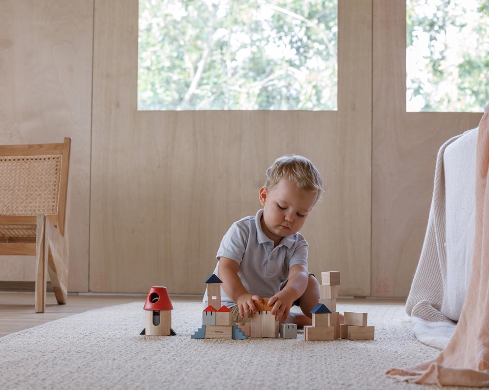 A child playing with the PlanToys Creative Blocks in the Orchard colour way. The child is sitting on a rug.  