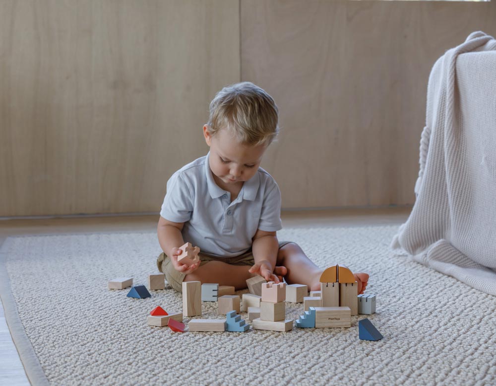 A child playing with the PlanToys Creative Blocks in the Orchard colour way. The child is sitting on a rug.  