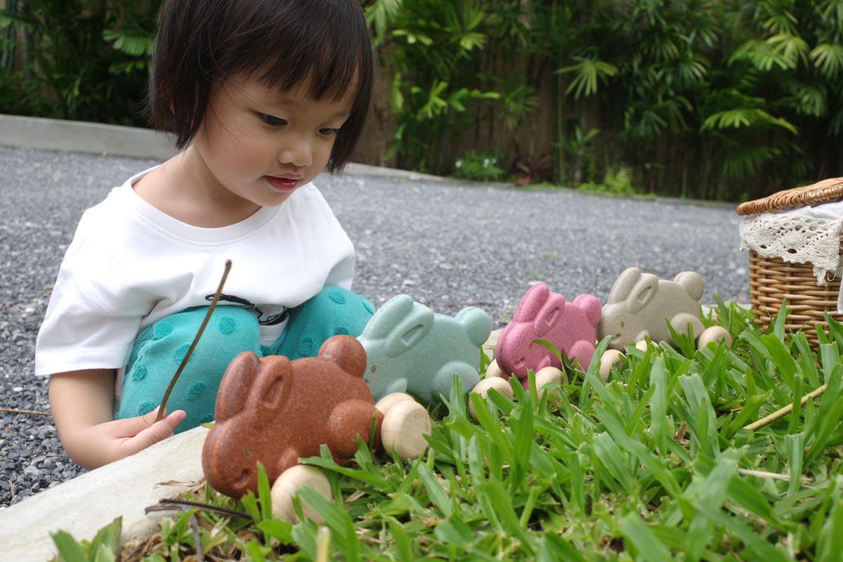 Girl crouched down looking at a line of Plan Toys plastic free rolling bunny toys lined up on some green grass