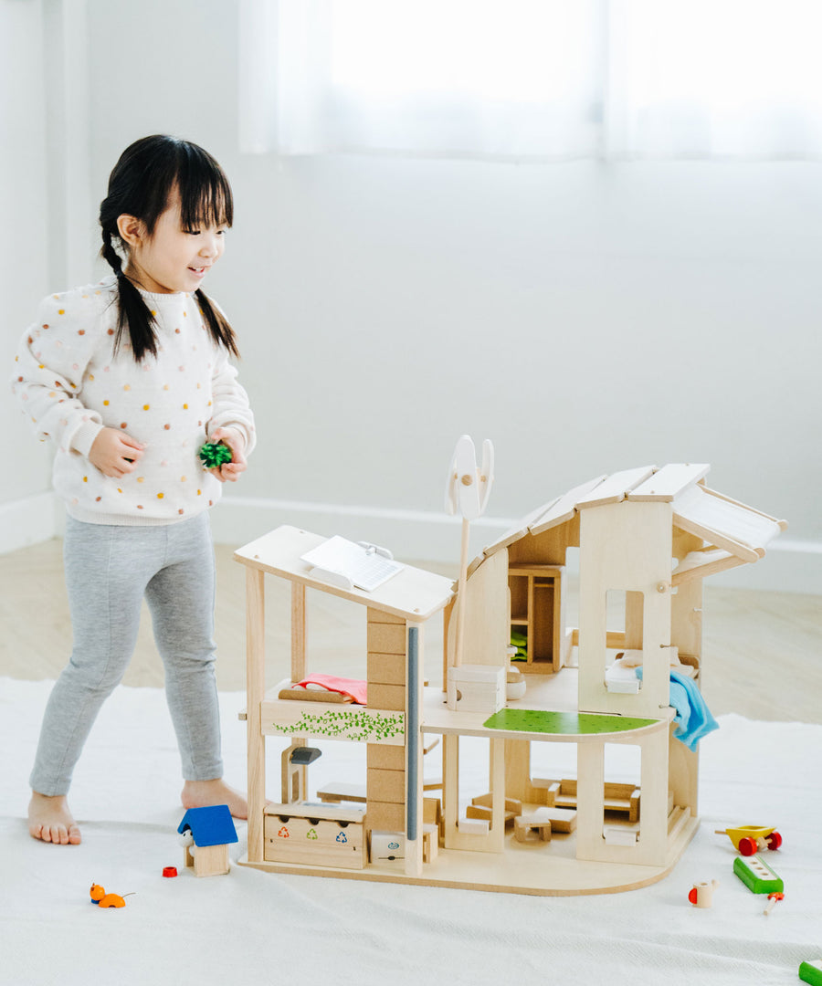 A child standing besides the PlanToys Green Dolls' House and Furniture Set. This eco inspired house has a lot of earth friendly features like a pretend recycling point, wind turbine, solar panel and grass and plants. 