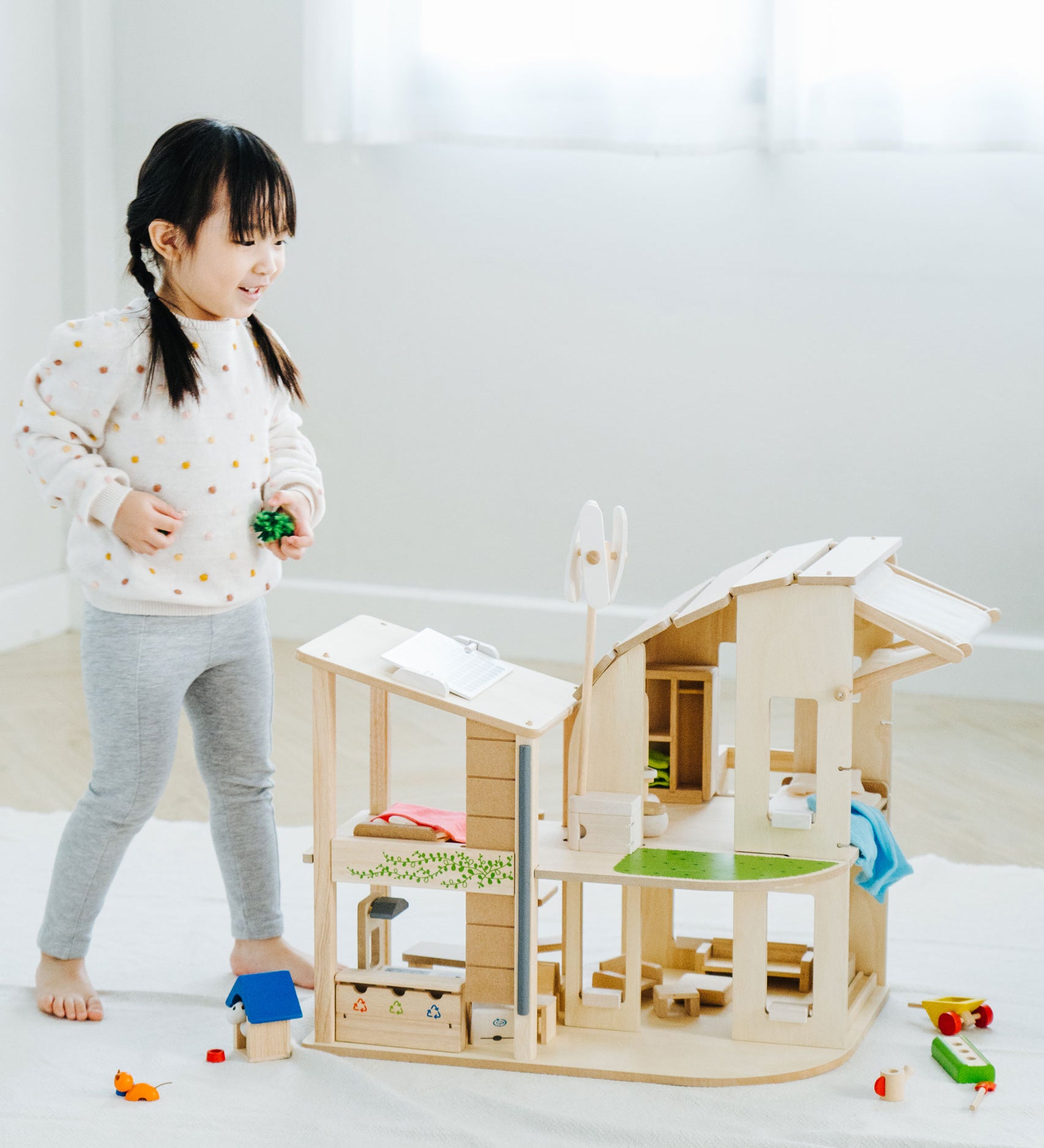 A child standing besides the PlanToys Green Dolls' House and Furniture Set. This eco inspired house has a lot of earth friendly features like a pretend recycling point, wind turbine, solar panel and grass and plants. 