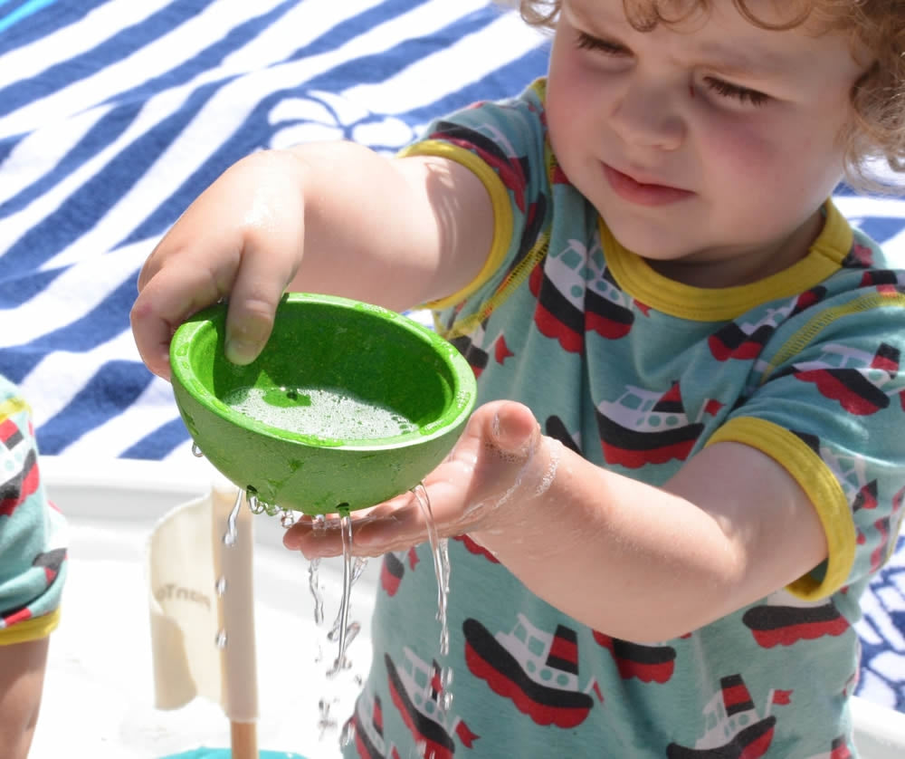 A child playing with the green bowl from the PlanToys Fountain Bowl Set, water can be seen pouring from the holes in side of the bowl.  