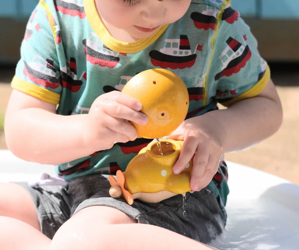 A child playing with the small yellow bowl from the PlanToys Fountain Bowl Set they are pouring water into a yellow PlanToys submarine. 