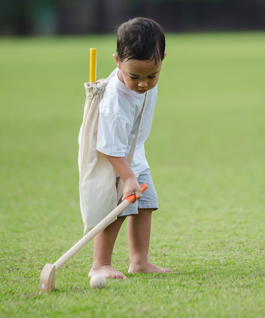 A child playing with a the PlanToys Mini Golf Set outside on grass. He is holding one of the clubs taking a swing at one of the balls. He has the canvas storage bag on his back with the other club poking out.