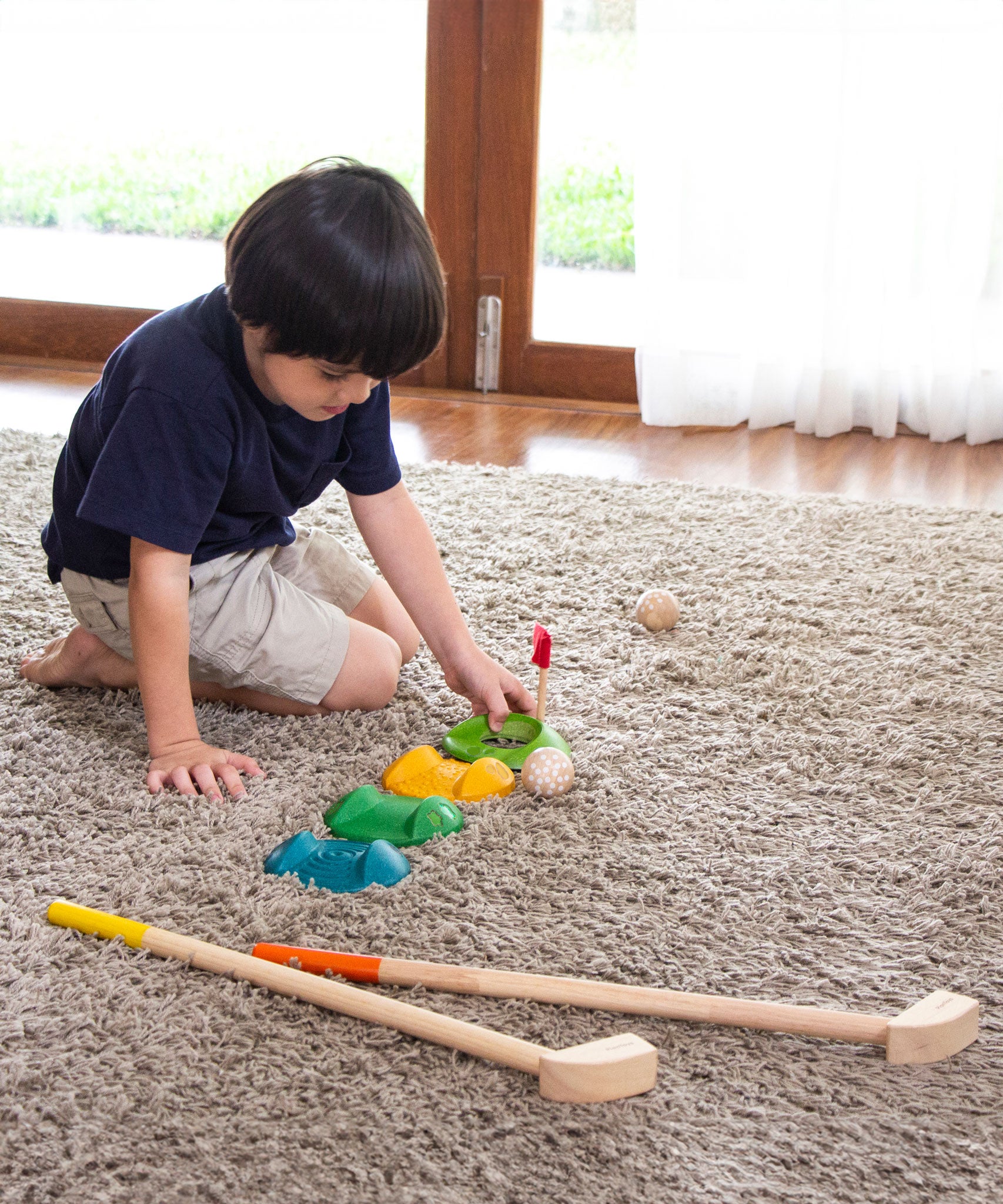 A child sitting on a rug setting up the pieces from the PlanToys mini wooden golf set.  