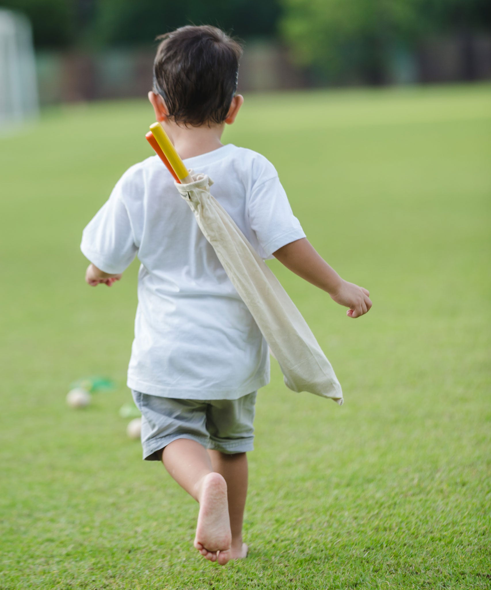 A child playing with a the PlanToys Mini Golf Set outside on grass. He is running towards the ball on the ground with the canvas bag containing the clubs on his back. 