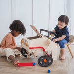 Two children playing with the PlanToys Motor Mechanic role play toy. One is sitting in front of the steering wheel and the other is fixing one of the wheels. 