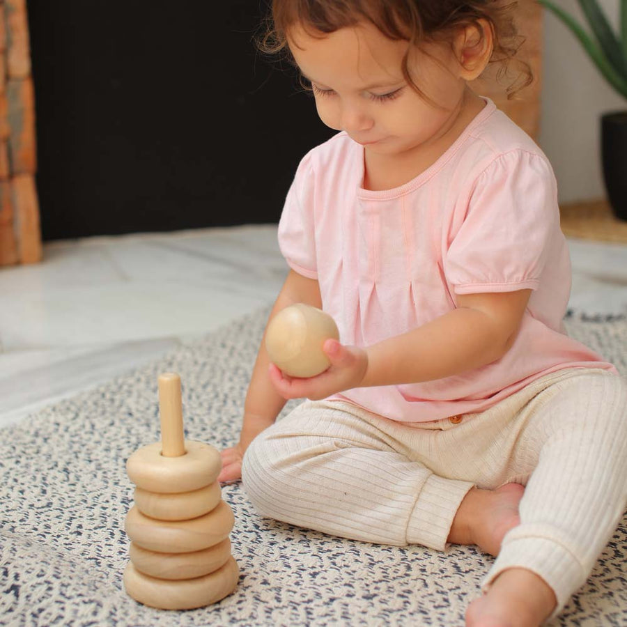 A child sitting on a blue and white rug playing with the Plan Toys Natural Stacking Ring. The child is holding the top wooden ball piece in their hand 