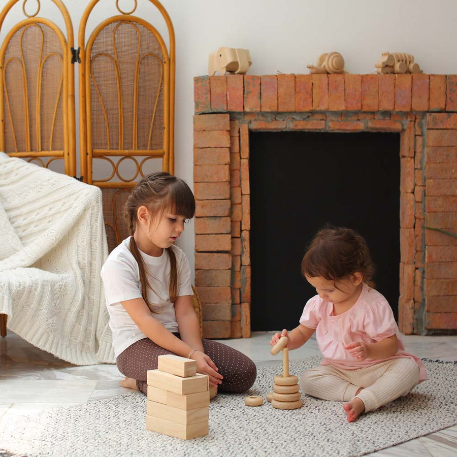 Two children sitting on a blue and white rug playing with the Plan Toys Natural Stacking Ring. The younger child is placing a ring piece onto the base. 