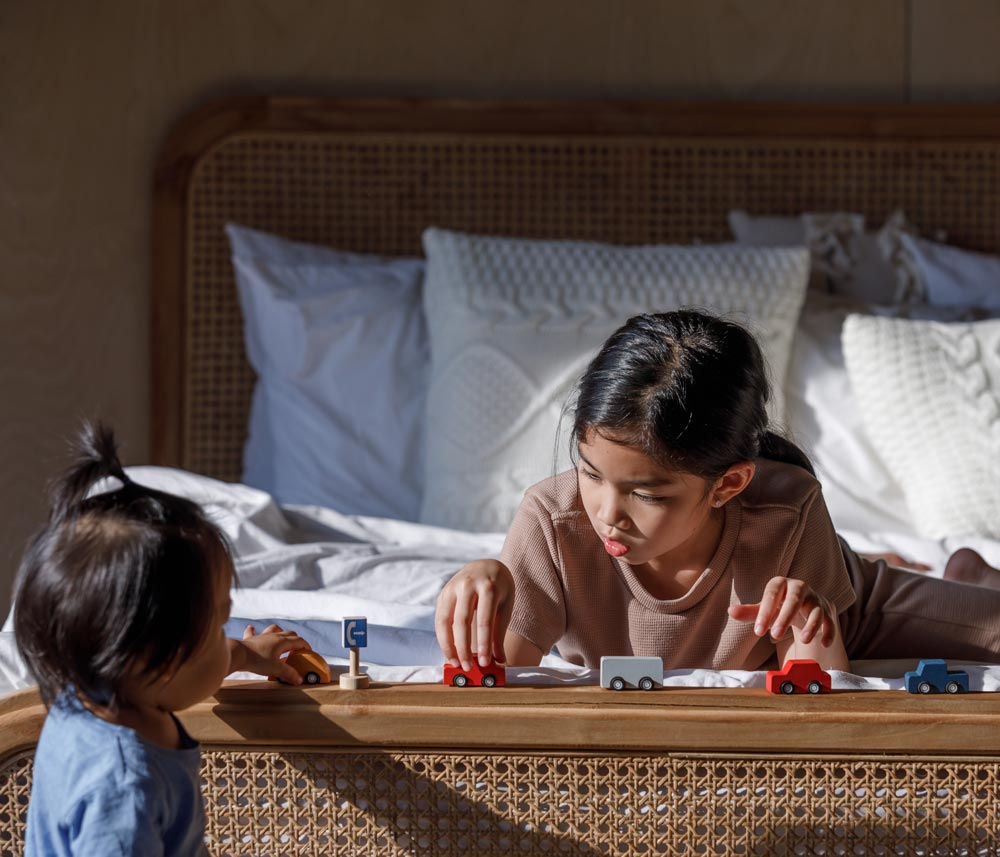 Two children playing with the PlanToys Mini Cars Set. One child is laying on a bed the other is standing up at the end of the bed.
