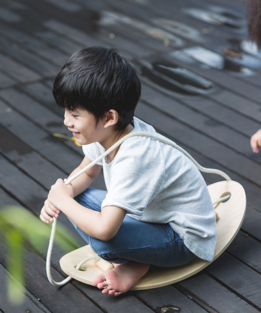 A child sitting on the PlanToys Balance Board holding the rope that is attached to the board. 