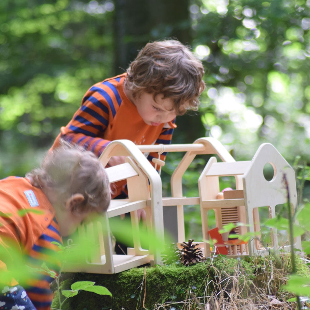 Children playing with the PlanToys Slide N Go Dolls' House placed on a moss covered tree stump in the forest. 