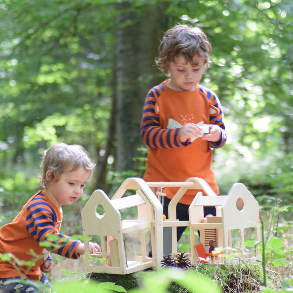 Children playing with the PlanToys Slide N Go Dolls' House that has been placed on a low tree stump in a forest. 
