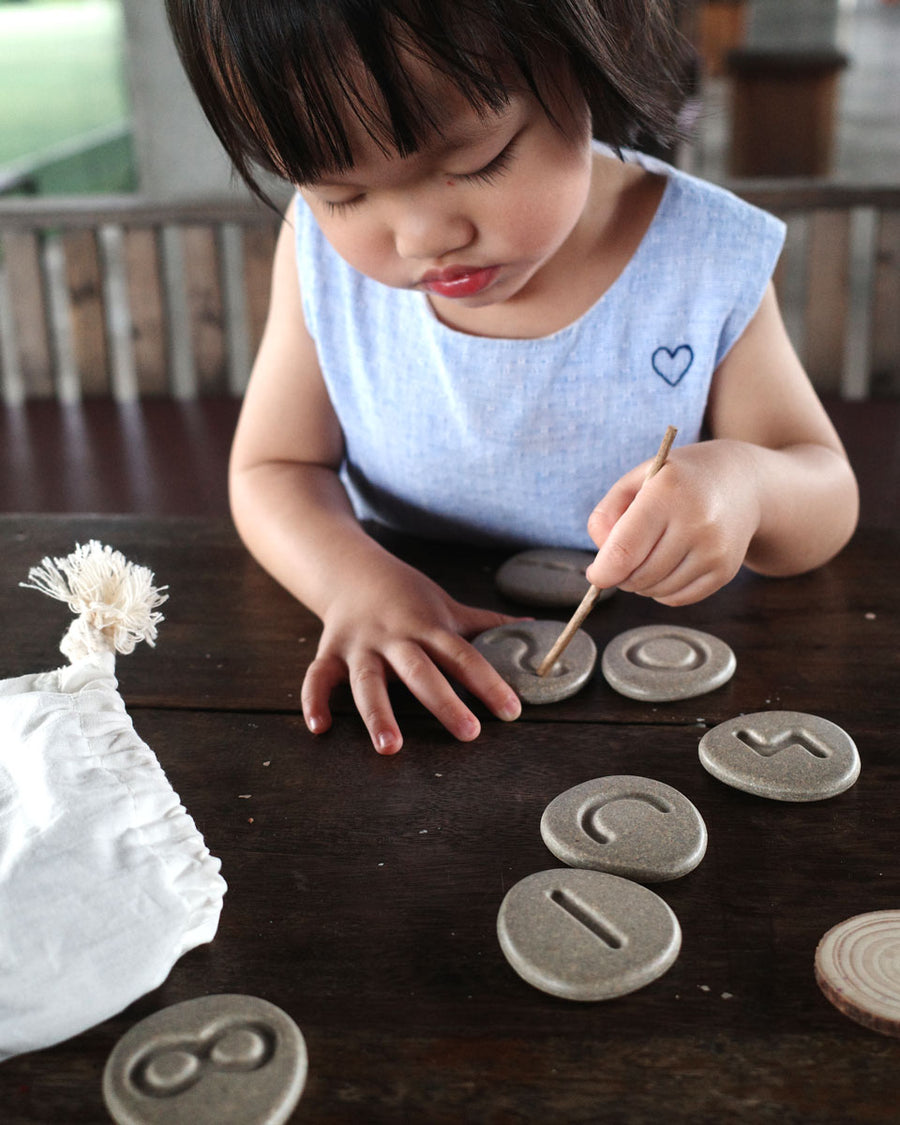 Cartoon hand reaching into a white drawstring bag filled with the Plan Toys eco-friendly wooden stone discs on a white background