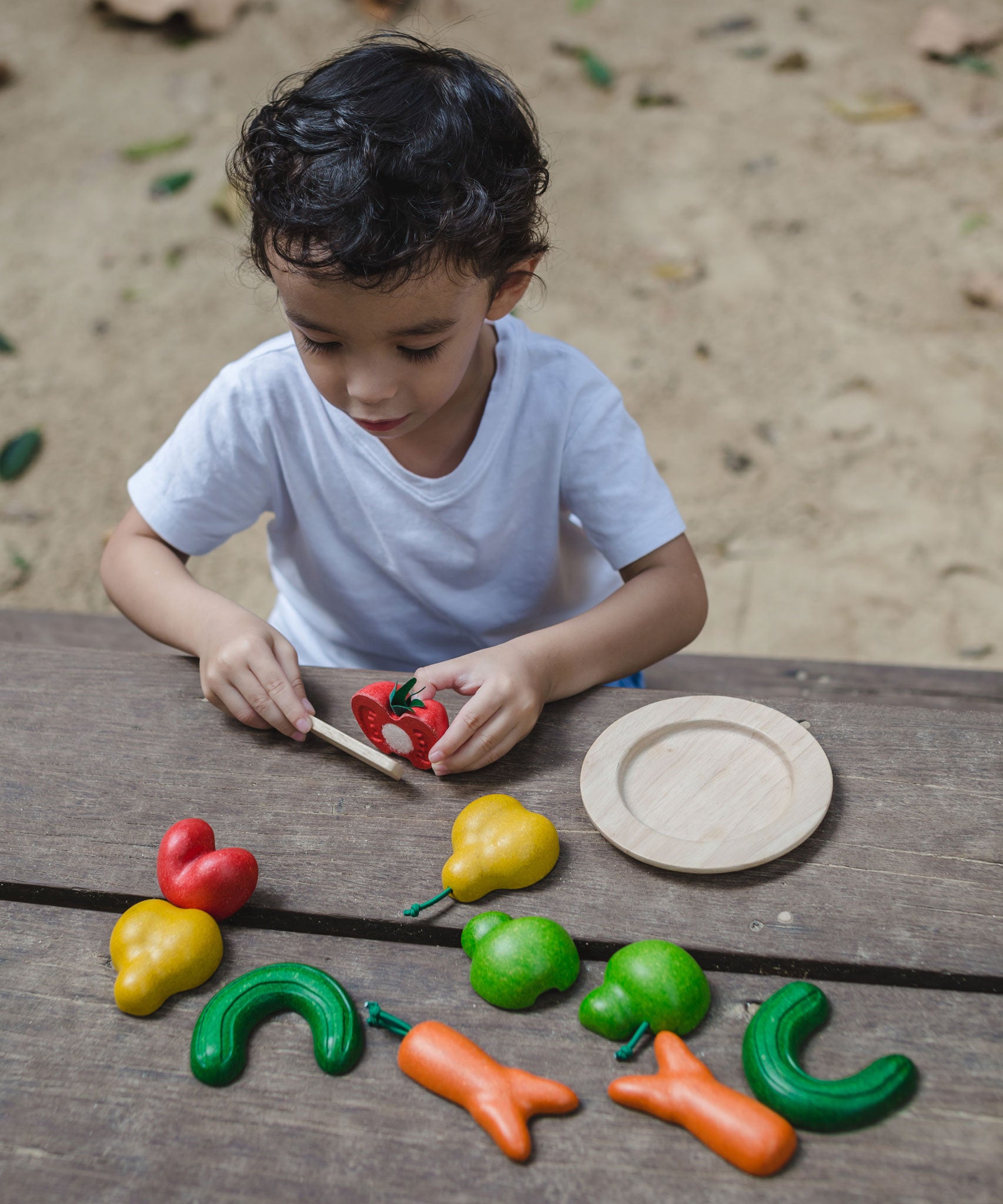 The PlanToys Wonky Fruit & Vegetables being played with by a child, the child is cutting the tomato in half using the wooden knife that is included. 