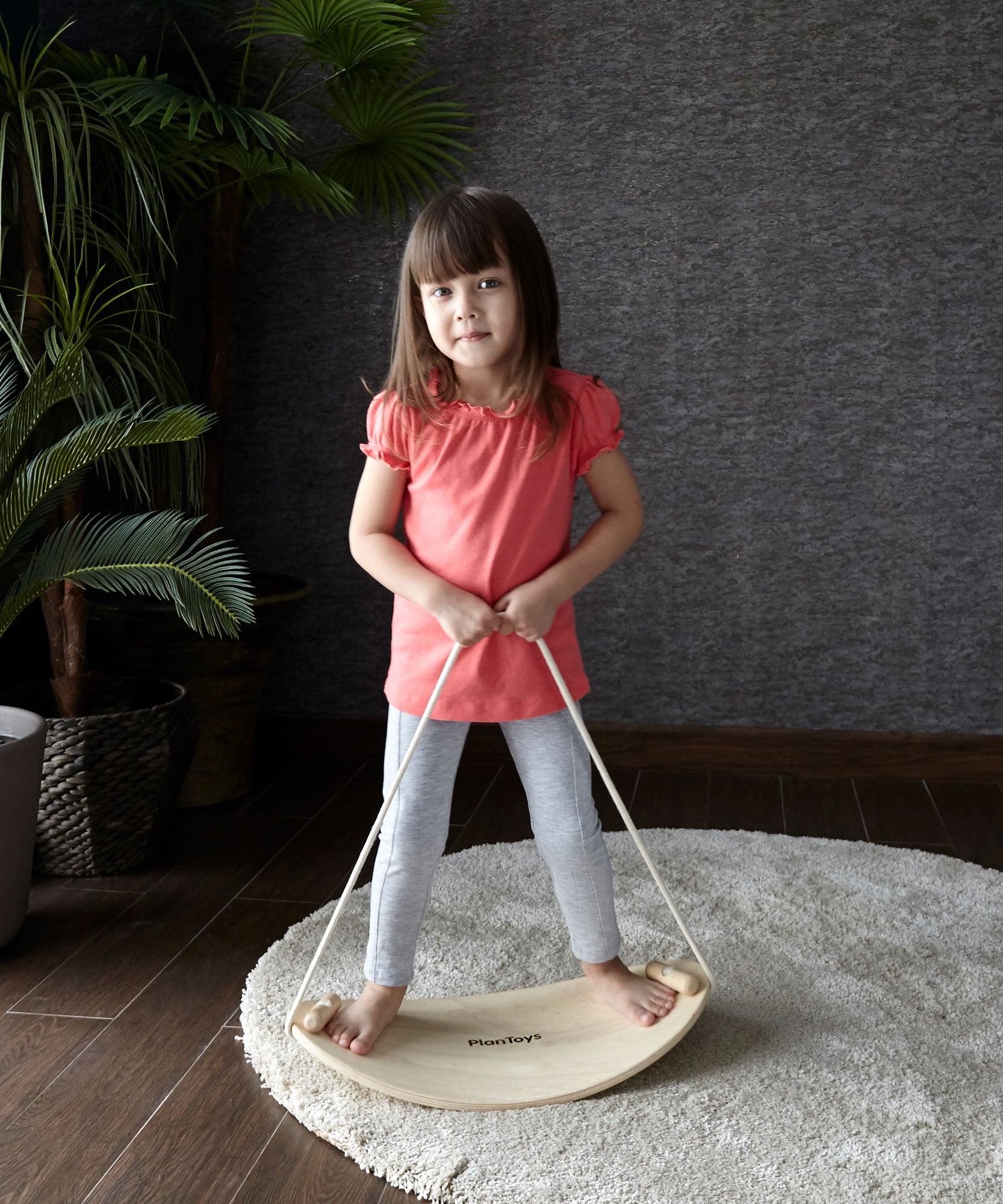 A child on the PlanToys Balance Board holding onto the rope attached to both sides of the board. The board has been placed on a circular shaped beige coloured rug. 