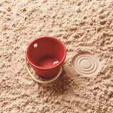 The PlanToys red bucket is stood on soft sane, the bucket is empty and the pattern on the base is imprinted on the sand. The pattern is 3 circles are in a bulls-eye pattern