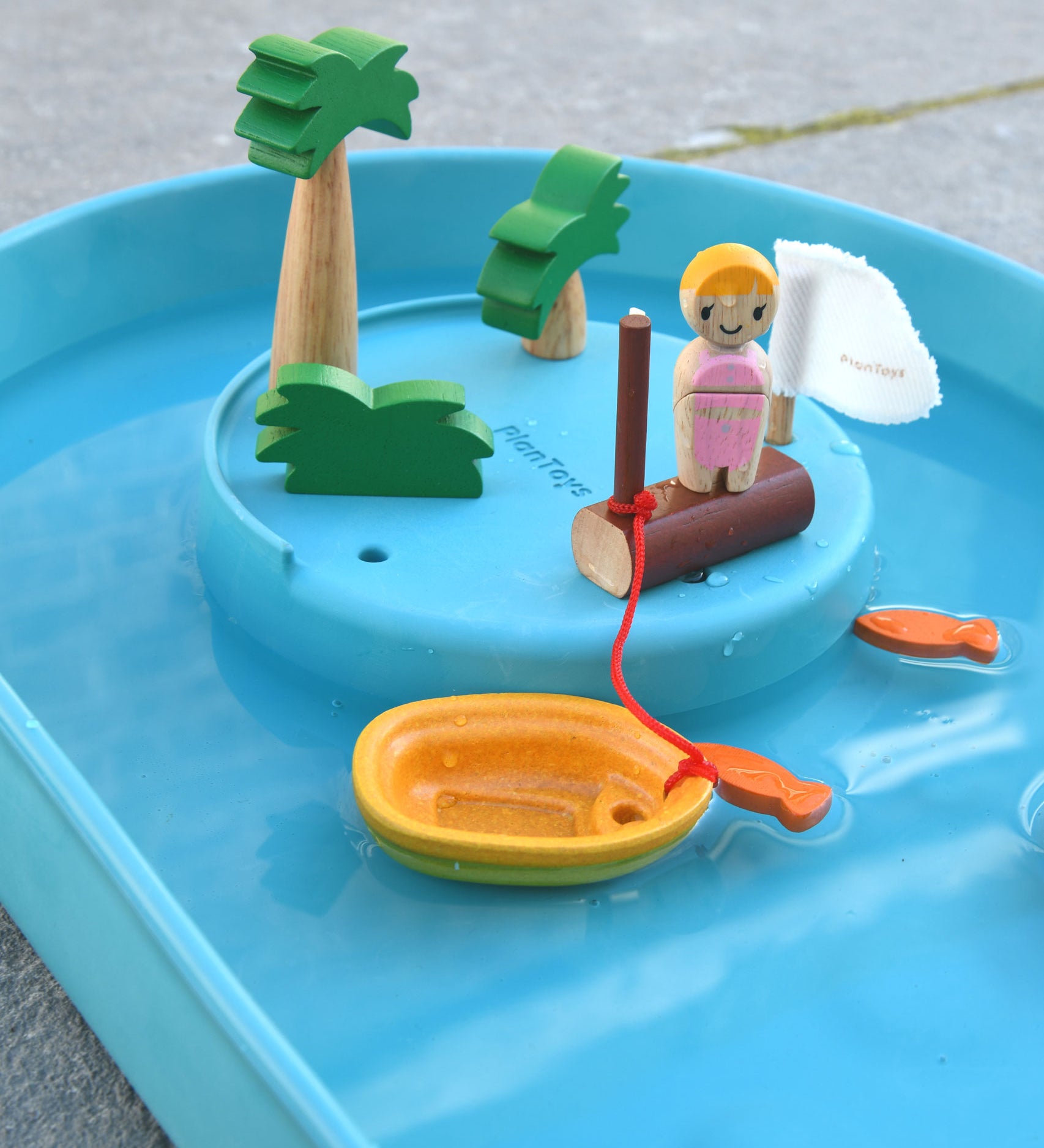 A close up of the raised section of the PlanToys Water Play Set. The wooden tree and doll figures have been placed on the raised section. The yellow boat has been anchored to the wooden post. Two orange fish can be seen in the water.