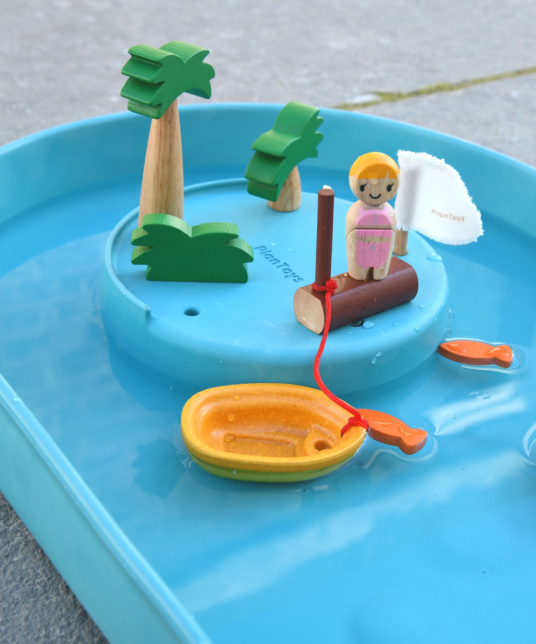 A close up of the raised section of the PlanToys Water Play Set. The wooden tree and doll figures have been placed on the raised section. The yellow boat has been anchored to the wooden post. Two orange fish can be seen in the water.