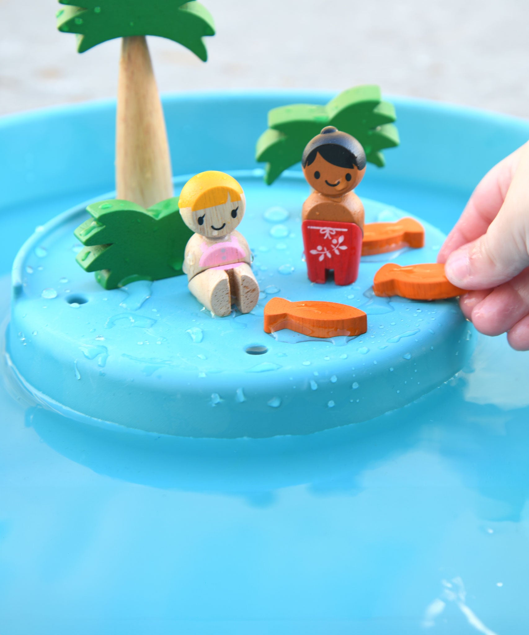 A close up of the raised 'island' section of the PlanToys Water Play Set. The two dollies are on the island surrounded by the trees and greenery and three orange fish. 