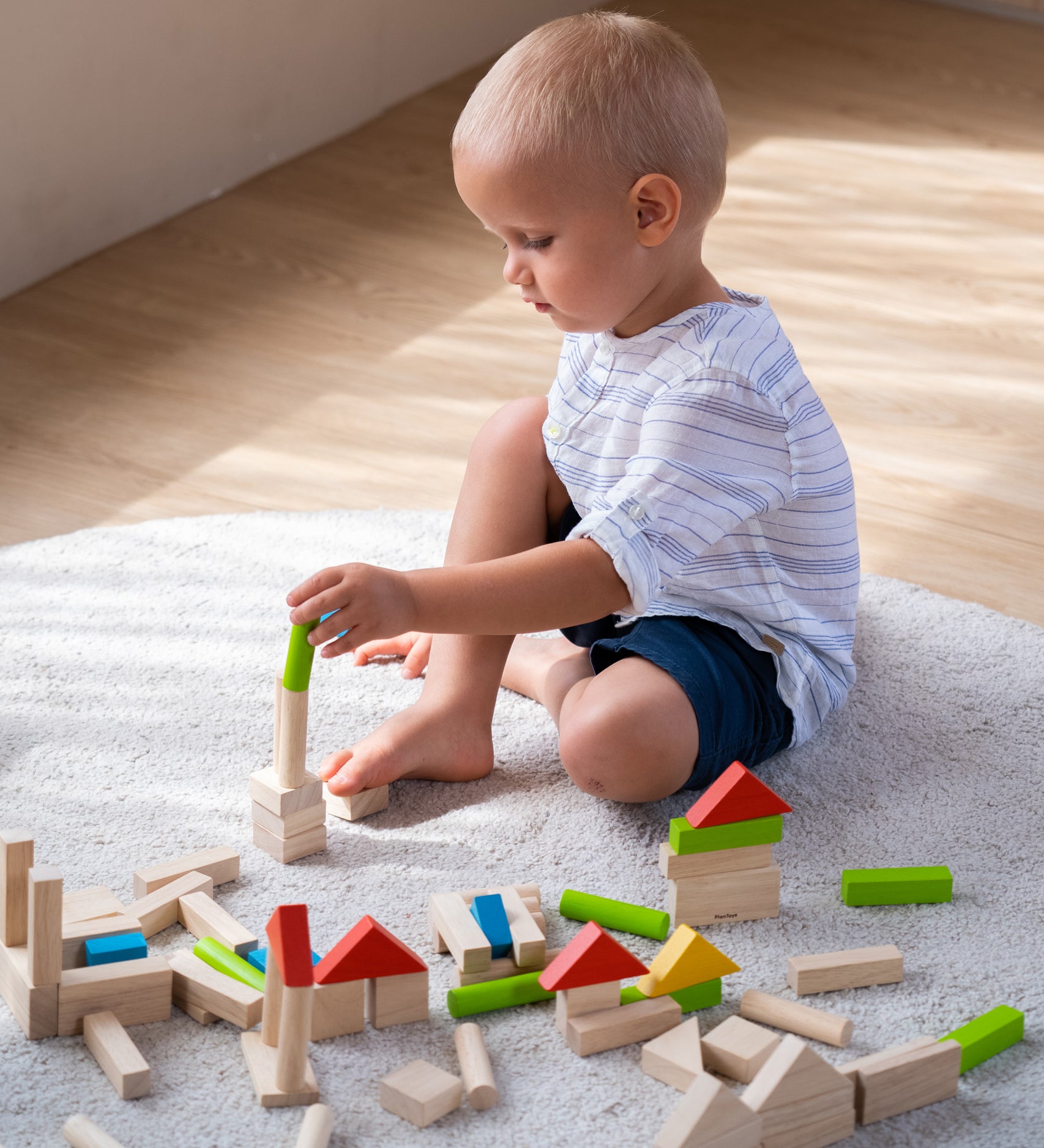 A child sat on a light grey coloured rug playing with the PlanToys 40 unit Colourful Block set. 
