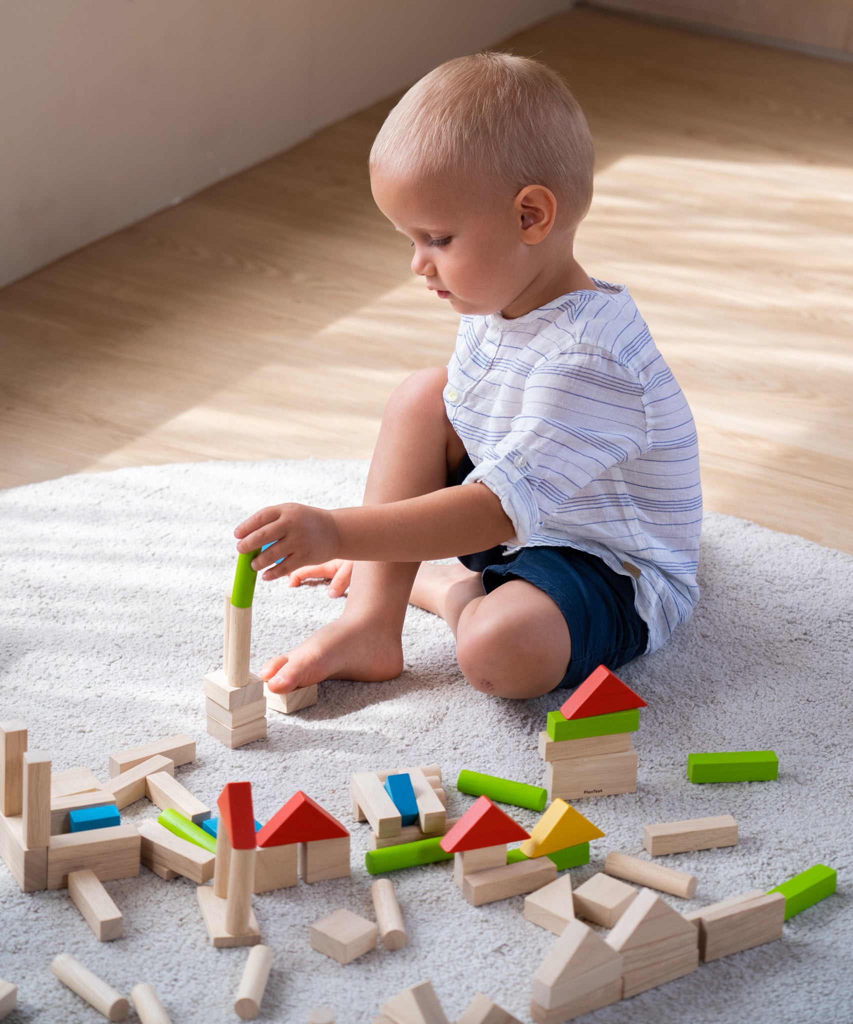 A child sat on a light grey coloured rug playing with the PlanToys 40 unit Colourful Block set. 
