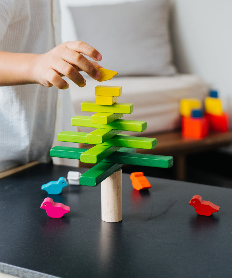 A close up of a child's hand placing the yellow coloured bird figures on the top of the Plan Toys Balancing Tree.
