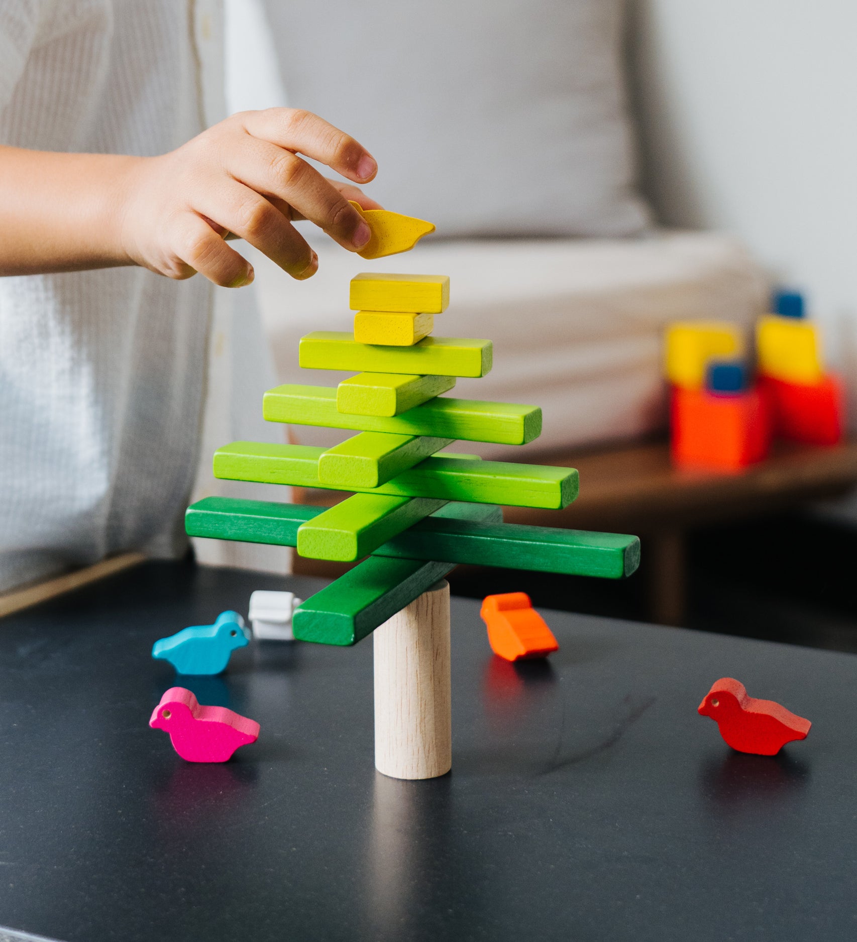 A close up of a child's hand placing the yellow coloured bird figures on the top of the Plan Toys Balancing Tree.
