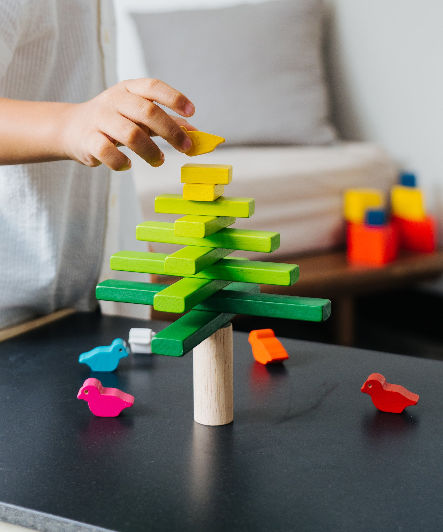 A close up of a child's hand placing the yellow coloured bird figures on the top of the Plan Toys Balancing Tree.
