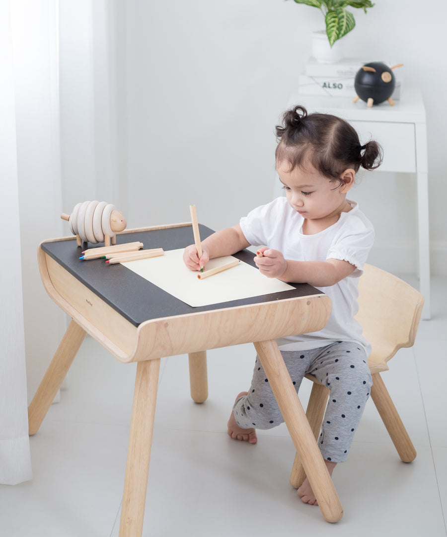 A child sitting by a PlanToys Black Table & Chair. The child is writing and a piece of paper. 